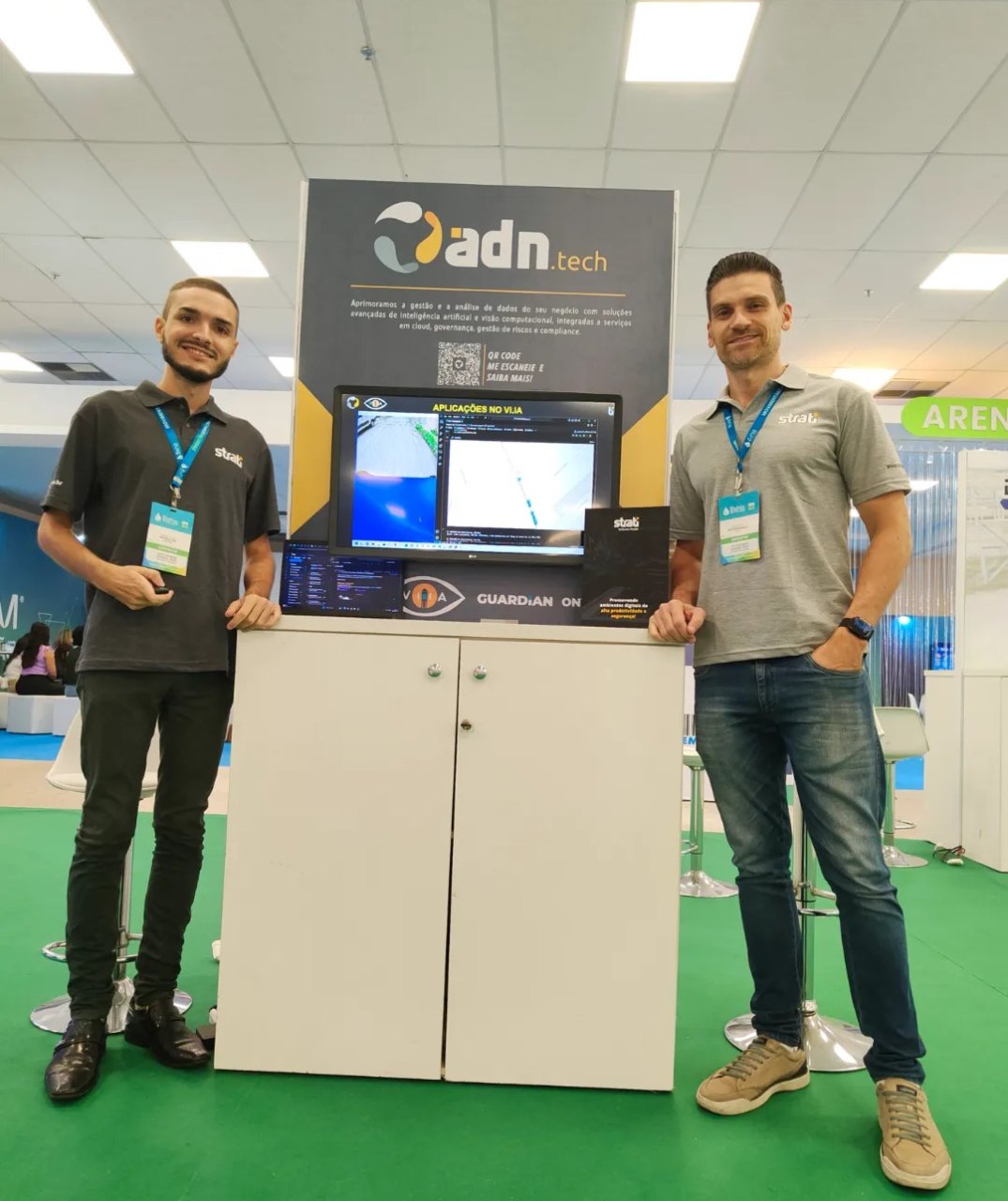 Matheus Ché e um colega no estande da ADN.tech, com um monitor e o logo da empresa