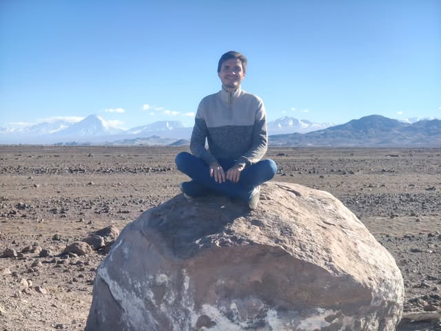 Matheus Ché sentado sobre uma pedra no deserto, com montanhas nevadas ao fundo